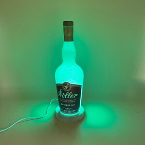 May include: A green illuminated bottle of Weller Antique 107 bourbon whiskey on a white marble base. The bottle is clear and has a textured surface. The base has a cord attached to it.