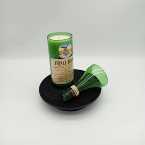 May include: A green glass candle holder with a white candle inside. The candle holder is shaped like a bottle and has the text "Fernet-Branca" on the label. The candle holder is sitting on a black circular base.