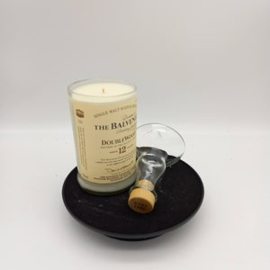 May include: A white candle in a clear glass bottle with a label that reads "The Balvenie Doublewood 12 Years Old" on a black circular base. A clear glass bottle with a cork stopper is next to the candle.