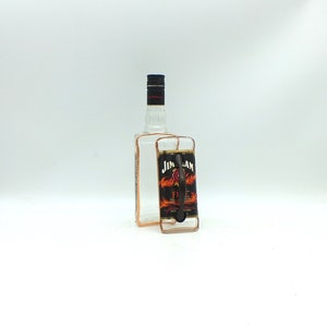 May include: A clear glass bottle with a black cap and a copper wire frame. A glass handle with a black label that reads "Jim Beam Fire" is attached to the bottle. The label features flames.