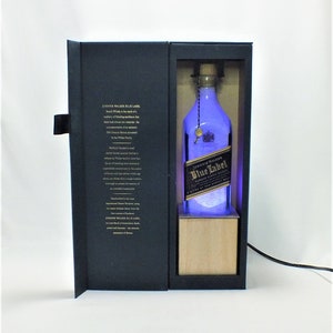 May include: A black box with a clear glass bottle of Johnnie Walker Blue Label Scotch Whisky inside. The bottle is illuminated with blue light and sits on a wooden base. The box has a gold inscription that reads "Johnnie Walker Blue Label" and a description of the whisky.
