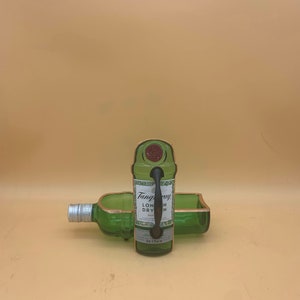 May include: A green glass bottle repurposed into a serving tray and a bottle holder. The bottle holder has a handle and the words "Tanqueray London Dry Gin" are visible on the label. The edges are trimmed with a copper-colored material.