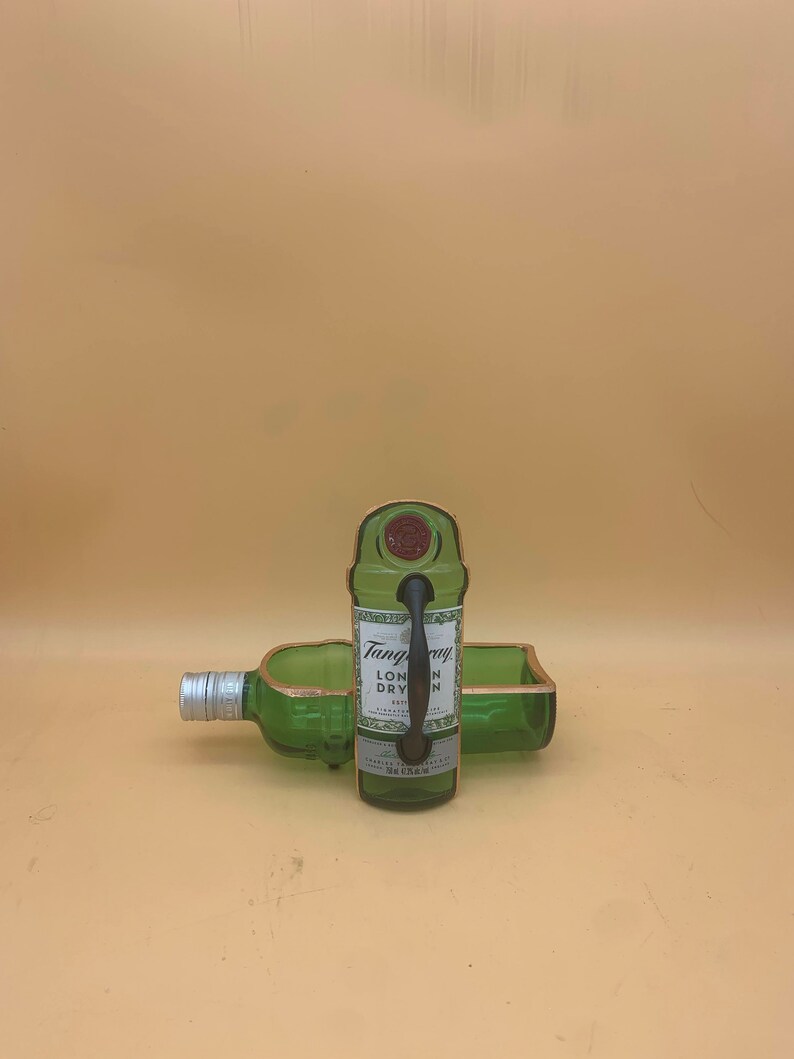 May include: A green glass bottle repurposed into a serving tray and a bottle holder. The bottle holder has a handle and the words "Tanqueray London Dry Gin" are visible on the label. The edges are trimmed with a copper-colored material.