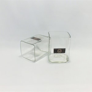 May include: Two clear glass square drinking glasses. The glass on the right has a black label with the number "12" printed on it.