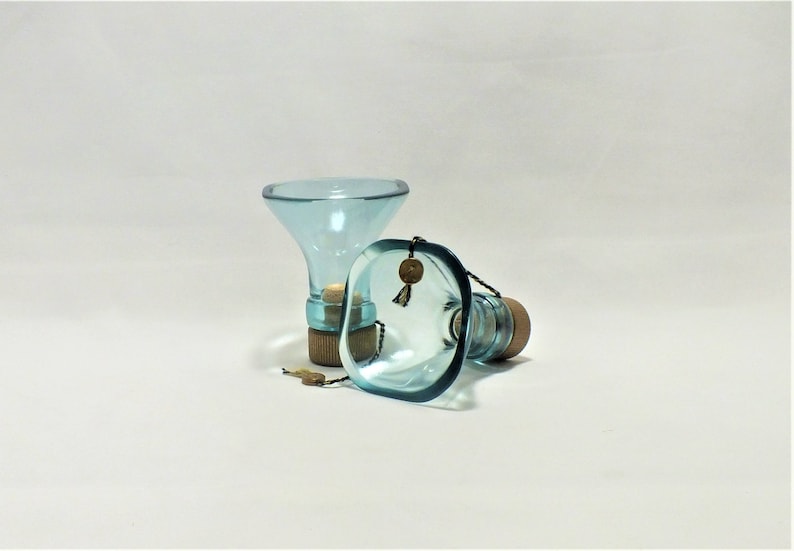 May include: Two clear blue glass bottle stoppers with cork tops. The stoppers are shaped like funnels and have a square base.