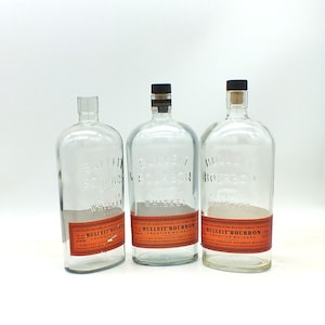 May include: Three clear glass Bulleit Bourbon whiskey bottles with red labels. The bottles are empty and have different types of stoppers. The labels read "Bulleit Bourbon Frontier Whiskey Kentucky Straight Bourbon Whiskey."