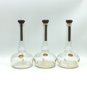 May include: Three clear glass bottles with long necks and rounded bases. Each bottle has a dark brown stopper and a gold-colored seal. A black and gold label is affixed to the neck of each bottle.