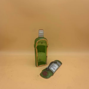 May include: A green glass bottle with a gold-colored trim and a silver cap. The bottle is cut in half, with the label lying next to it. The label reads "Tanqueray London Dry Gin".