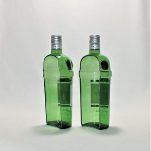 Op de afbeelding: Twee lege groene glazen gin flessen met zilveren doppen. De flessen zijn licht gebogen en hebben een etiket aan de voorkant.