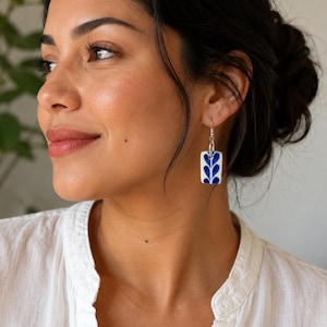 May include: White and blue rectangular earrings with a floral design. The earrings are hanging from silver hooks. The blue design is a stylized leaf pattern. The earrings are a unique accessory.