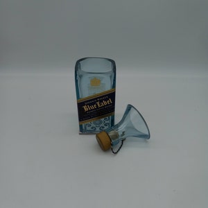 May include: A clear glass bottle with a blue label that reads "Johnnie Walker Blue Label Blended Scotch Whisky" and a gold cap. The bottle is sitting on a white surface. A small blue glass funnel with a gold cap is next to the bottle.