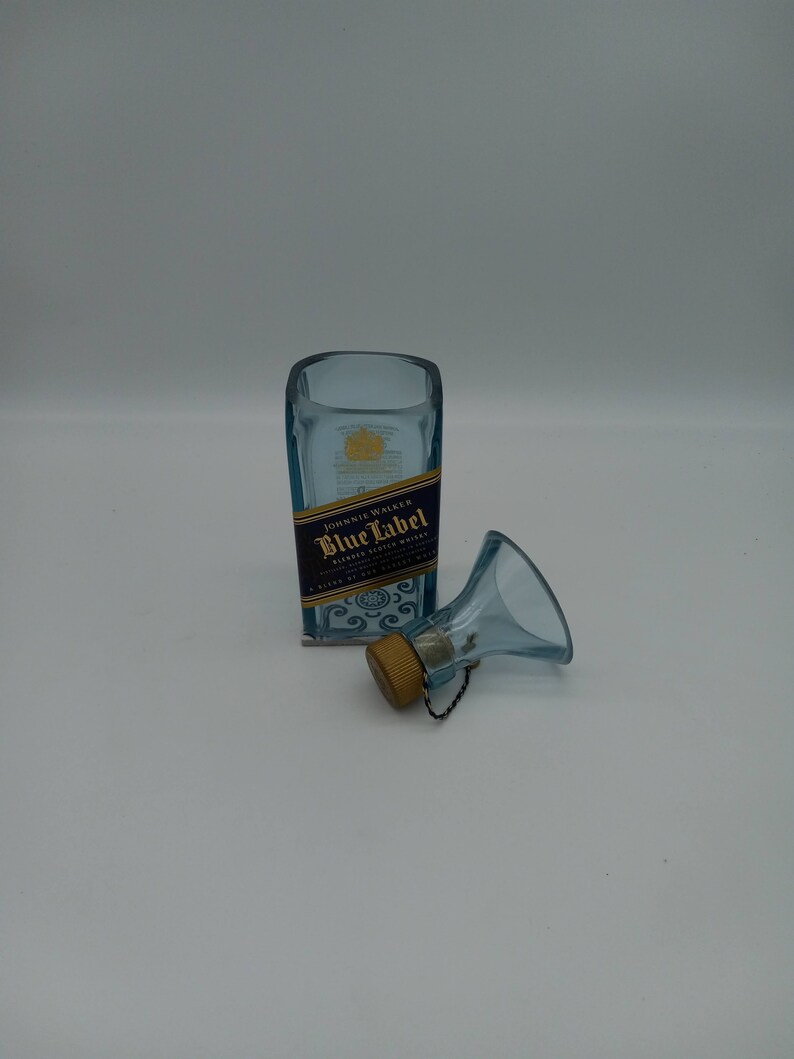 May include: A clear glass bottle with a blue label that reads "Johnnie Walker Blue Label Blended Scotch Whisky" and a gold cap. The bottle is sitting on a white surface. A small blue glass funnel with a gold cap is next to the bottle.
