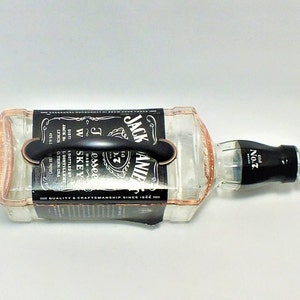 May include: A clear glass bottle of Jack Daniel's Tennessee Whiskey, with a black label and handle. The label features the brand name and other text. The bottle is lying on its side.