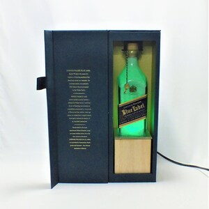May include: A blue and gold box with a bottle of Johnnie Walker Blue Label Scotch Whisky inside. The bottle is illuminated with a green light. The box has a description of the whisky on the front.