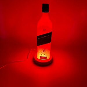 Pode incluir: Uma luz vermelha ilumina uma garrafa de vidro transparente com um r&oacute;tulo preto que diz "JOHNNIE WALKER. BLACK LABEL. 12" e uma silhueta de veado. A garrafa est&aacute; sobre uma base branca com um cabo.