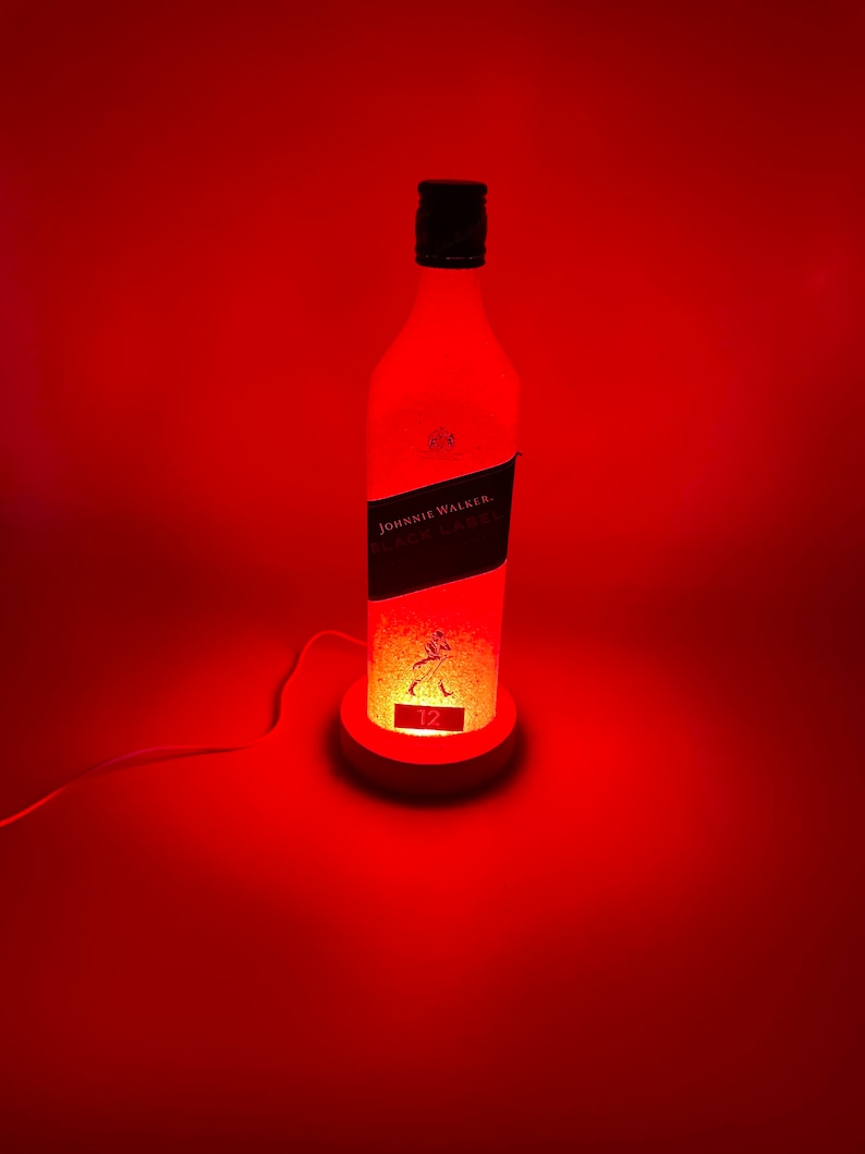 Pode incluir: Uma luz vermelha ilumina uma garrafa de vidro transparente com um r&oacute;tulo preto que diz "JOHNNIE WALKER. BLACK LABEL. 12" e uma silhueta de veado. A garrafa est&aacute; sobre uma base branca com um cabo.