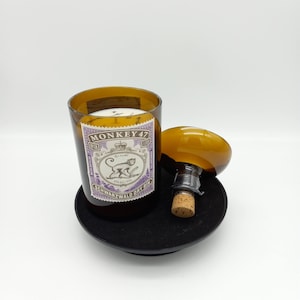 May include: A brown glass candle jar with a white wax candle inside. The jar has a label with the text "MONKEY 47" and a monkey illustration. The jar is sitting on a black coaster with a brown bottle stopper.