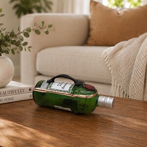 May include: A green wine bottle with a black handle and a silver stopper rests on a wooden coffee table. The bottle has a label and a red wax seal. The background includes a beige sofa, a throw blanket, and a decorative vase.