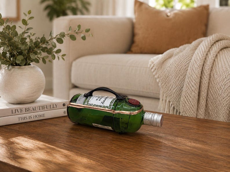 May include: A green wine bottle with a black handle and a silver stopper rests on a wooden coffee table. The bottle has a label and a red wax seal. The background includes a beige sofa, a throw blanket, and a decorative vase.