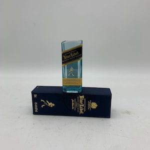May include: A miniature glass bottle with a blue label that reads "Johnnie Walker Blue Label Blended Scotch Whisky". The bottle is sitting on top of a blue box with the same label.