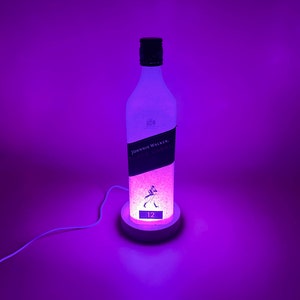 Pode incluir: Uma garrafa de vidro transparente com um r&oacute;tulo branco que diz "Johnnie Walker Black Label 12" est&aacute; iluminada com luz roxa. A garrafa est&aacute; sentada em uma base branca com um cabo.