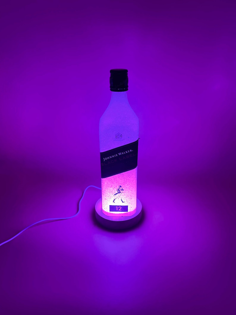 Pode incluir: Uma garrafa de vidro transparente com um r&oacute;tulo branco que diz "Johnnie Walker Black Label 12" est&aacute; iluminada com luz roxa. A garrafa est&aacute; sentada em uma base branca com um cabo.
