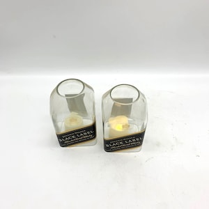 May include: Two clear glass candle holders shaped like Johnnie Walker Black Label whiskey bottles. The bottles have gold labels with black text that reads "Johnnie Walker Black Label Blended Scotch Whisky".