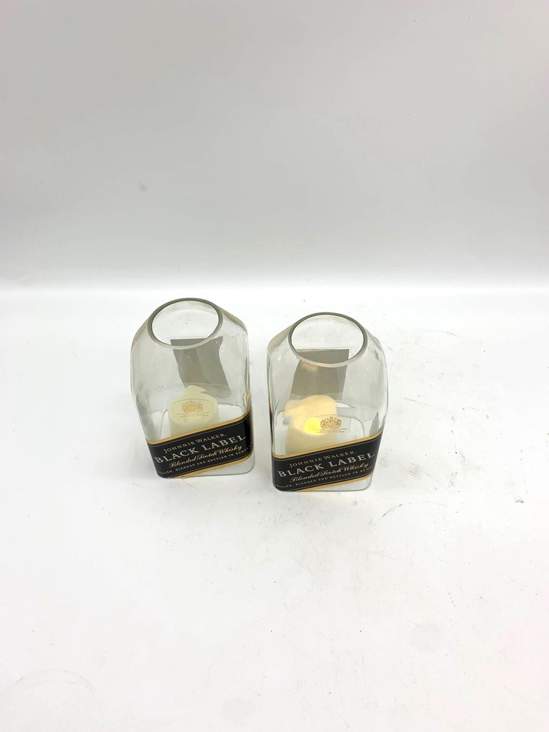 May include: Two clear glass candle holders shaped like Johnnie Walker Black Label whiskey bottles. The bottles have gold labels with black text that reads "Johnnie Walker Black Label Blended Scotch Whisky".