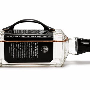May include: A clear glass Jack Daniel's bottle with a black handle and a black label. The bottle has a copper-colored wire wrapped around it. The label has white text and a picture of a man.