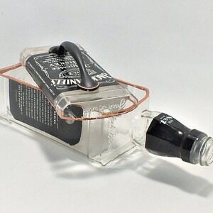 May include: A repurposed Jack Daniel's whiskey bottle, transformed into a unique container. The clear glass bottle features a black label and a copper-colored frame, with a black handle attached to the top. The bottle is lying on its side.