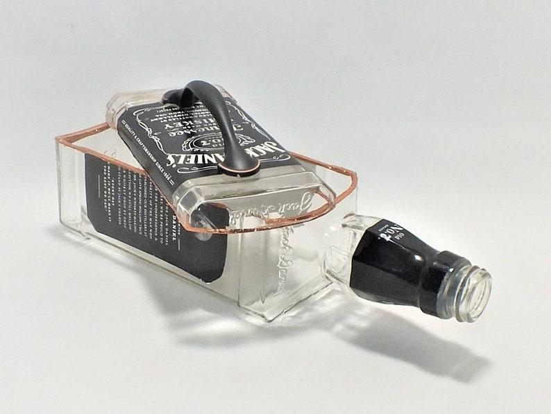 May include: A repurposed Jack Daniel's whiskey bottle, transformed into a unique container. The clear glass bottle features a black label and a copper-colored frame, with a black handle attached to the top. The bottle is lying on its side.