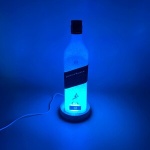 Pode incluir: Uma luz azul ilumina uma garrafa de vidro transparente com um r&oacute;tulo preto que diz "Johnnie Walker" e "12". A garrafa est&aacute; sentada em uma base branca com um cabo.