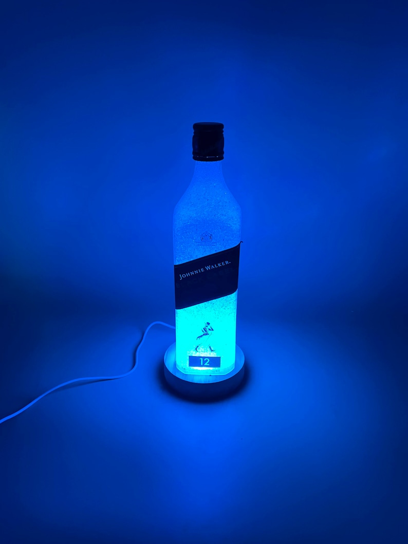 Pode incluir: Uma luz azul ilumina uma garrafa de vidro transparente com um r&oacute;tulo preto que diz "Johnnie Walker" e "12". A garrafa est&aacute; sentada em uma base branca com um cabo.