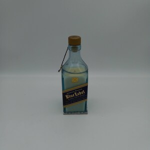May include: A clear glass bottle candle with a wooden cork top. The bottle is decorated with a blue and gold label that reads "Johnnie Walker Blue Label Blended Scotch Whisky".