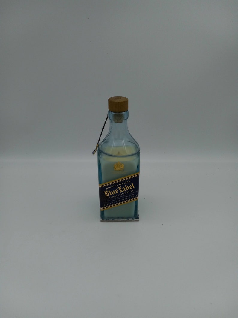 May include: A clear glass bottle candle with a wooden cork top. The bottle is decorated with a blue and gold label that reads "Johnnie Walker Blue Label Blended Scotch Whisky".