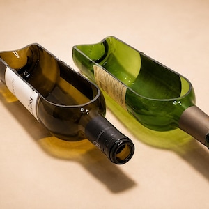 May include: Two glass wine bottles cut lengthwise to create open, trough-like containers. One bottle is dark green with a partial white label, and the other is a lighter green with a tan label. Both retain their original necks and bottle caps.