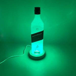Pode incluir: Uma garrafa de vidro transparente com um r&oacute;tulo preto que diz "JOHNNIE WALKER. BLACK LABEL." A garrafa &eacute; iluminada por dentro, projetando um brilho verde. A garrafa est&aacute; sentada em uma base branca com o n&uacute;mero "12" nela.