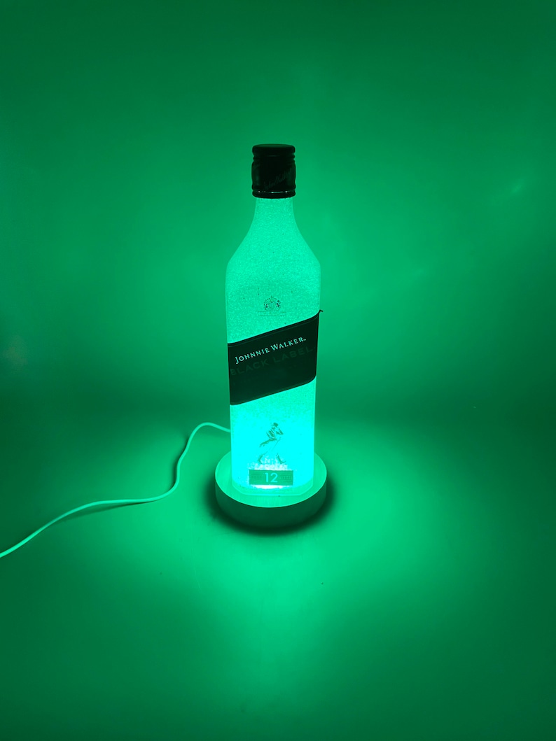 Pode incluir: Uma garrafa de vidro transparente com um r&oacute;tulo preto que diz "JOHNNIE WALKER. BLACK LABEL." A garrafa &eacute; iluminada por dentro, projetando um brilho verde. A garrafa est&aacute; sentada em uma base branca com o n&uacute;mero "12" nela.