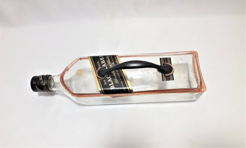 Puede incluir: Una bandeja de servicio hecha de una botella reciclada de Johnnie Walker Black Label. La bandeja de vidrio transparente tiene un borde de color cobre y un asa negra. La etiqueta original a&uacute;n es visible.