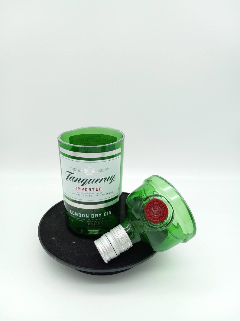 K&ouml;nnte beinhalten: Eine gr&uuml;ne Glasflasche Tanqueray London Dry Gin, mit abgenommenem Flaschenverschluss und der Flasche auf einem schwarzen runden Untersetzer. Die Flasche ist mit "Tanqueray Importiert" und "London Dry Gin" beschriftet.