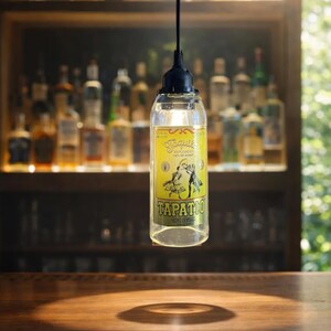May include: A hanging pendant light fixture fashioned from a clear glass Tapatio tequila bottle. The bottle features a yellow label with black text and a graphic illustration of two figures dancing. A black cord suspends the bottle from above.