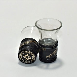May include: Two clear glass shot glasses with black plastic bases. The bases are decorated with the Johnnie Walker Black Label logo and the text "12 Years Old".