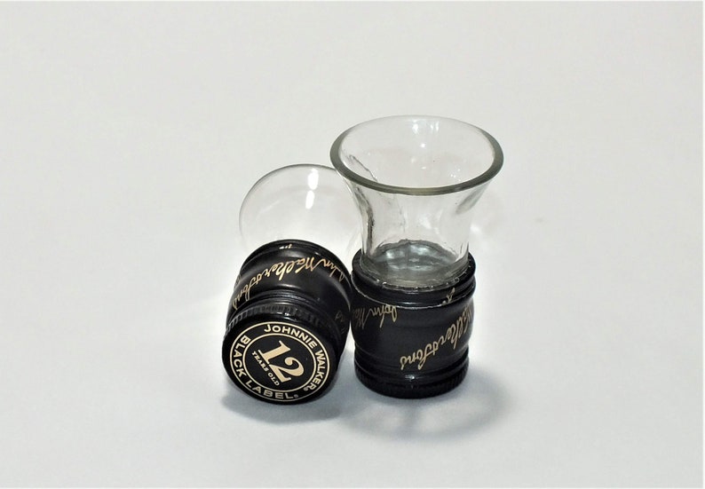 May include: Two clear glass shot glasses with black plastic bases. The bases are decorated with the Johnnie Walker Black Label logo and the text "12 Years Old".