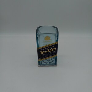May include: A clear glass bottle with a blue label that says "Johnnie Walker Blue Label Blended Scotch Whisky" and a gold crown on the top. The bottle is sitting on a white surface.
