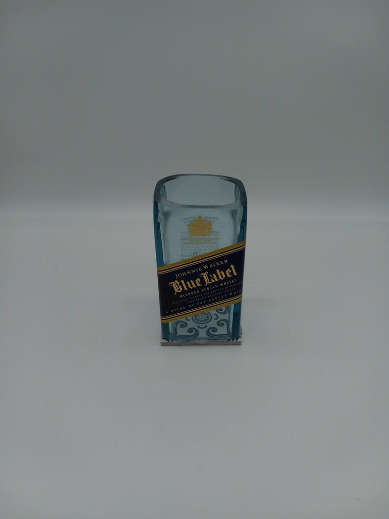 May include: A clear glass bottle with a blue label that says "Johnnie Walker Blue Label Blended Scotch Whisky" and a gold crown on the top. The bottle is sitting on a white surface.