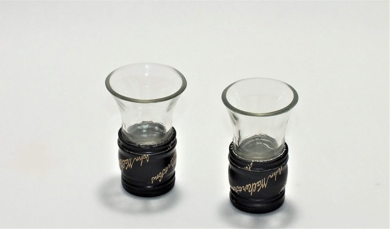 May include: Two clear glass shot glasses with black bases. The bases are wrapped with a black band with gold lettering that reads "Johnnie Walker" and "Red Label".