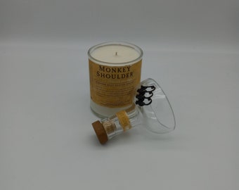 Könnte beinhalten: Eine weiße Kerze in einem durchsichtigen Glasgefäß mit einem Etikett, auf dem "Monkey Shoulder" Blended Malt Scotch Whisky steht. Die Kerze steht auf einer weißen Oberfläche.