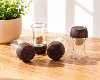 May include: Three bottle stoppers featuring clear glass tops and dark brown wooden bases with cork inserts. The wooden tops are rounded and smooth, with one displaying a visible engraved signature. The stoppers are arranged on a wooden surface.