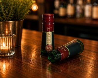 May include: Two small green glass bottles with dark red screw caps. One bottle stands upright, while the other lies on its side. The labels feature the text John Jameson, JJ&S, and a vertical code reading JO-0-585-CR in white and gold lettering.