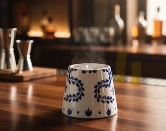 Könnte beinhalten: Ein weißer Keramikbecher mit blauem Blumendesign, gefüllt mit Eis und Dampf, steht auf einer Holzbar. Der Becher hat eine ausgestellte Form und ist mit blauen, blattartigen Mustern verziert. Barkeeper-Werkzeuge und Flaschen sind im Hintergrund zu sehen.